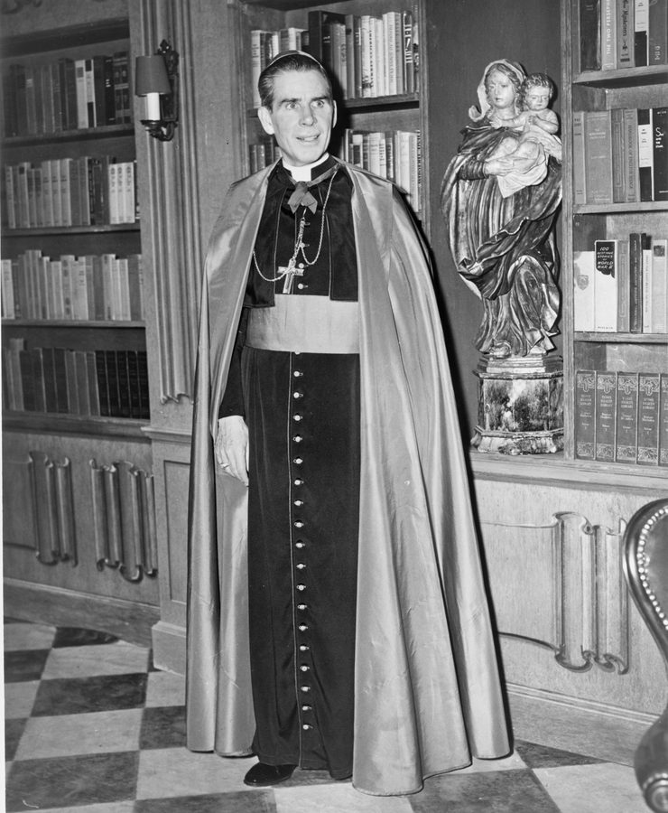 Bishop Fulton J. Sheen in cassock, ferraiolo and pectoral cross, standing among bookshelves beside a carved statue of the Madonna and Child, on the set of his television programme.